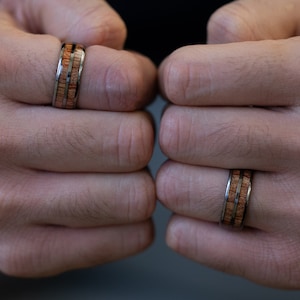 K&ouml;nnte beinhalten: Zwei Ringe aus Holz und Metall an den Fingern einer Person. Die Ringe haben eine braune Holzeinlage und eine silberne Metallband.