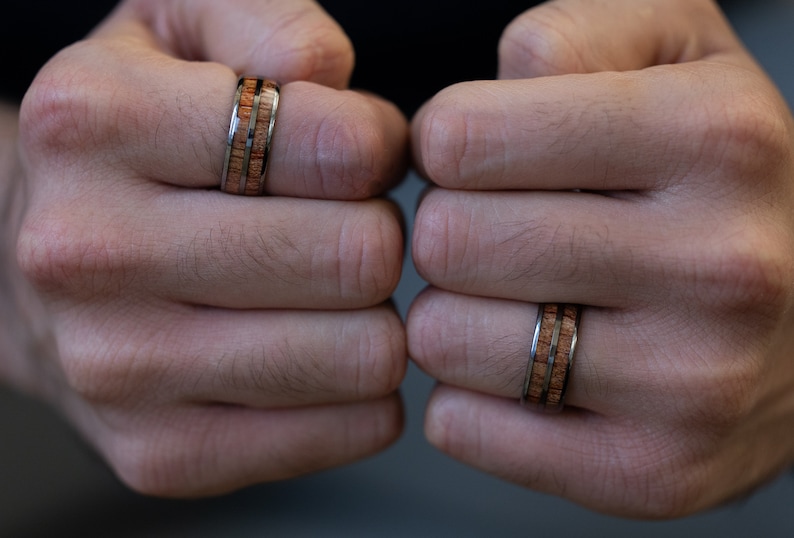 K&ouml;nnte beinhalten: Zwei Ringe aus Holz und Metall an den Fingern einer Person. Die Ringe haben eine braune Holzeinlage und eine silberne Metallband.