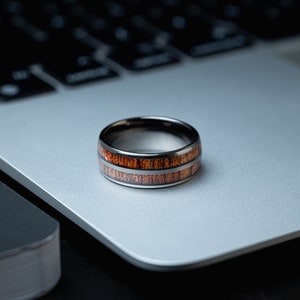 K&ouml;nnte beinhalten: Ein schwarzer und brauner Ring mit einem Holz-Inlay-Design. Der Ring liegt auf einem silbernen Laptop.