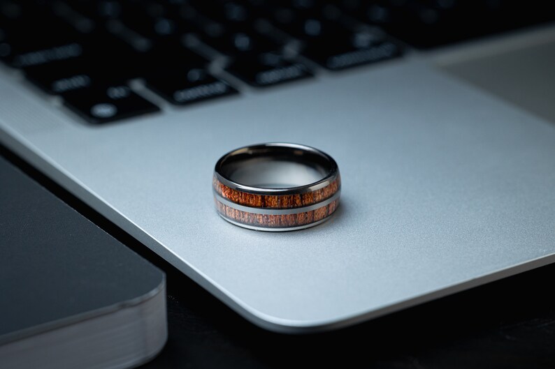 K&ouml;nnte beinhalten: Ein schwarzer und brauner Ring mit einem Holz-Inlay-Design. Der Ring liegt auf einem silbernen Laptop.