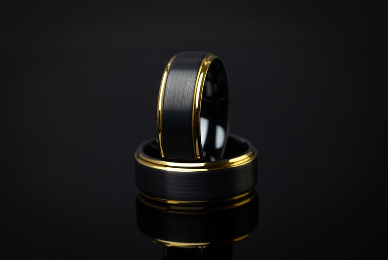 May include: Two black tungsten carbide rings with a gold band around the edge. The rings are stacked on top of each other.