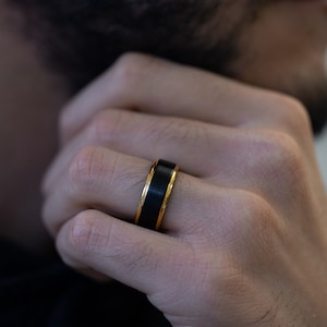 May include: A black ring with a gold band around the edge. The ring is worn on the finger of a person's hand.