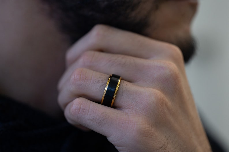May include: A black ring with a gold band around the edge. The ring is worn on the finger of a person's hand.