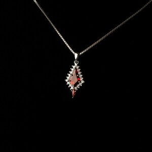 May include: A silver necklace with a pendant featuring a large, red gemstone surrounded by small, clear stones. The pendant has a unique, elongated shape. The background is solid black, highlighting the jewelry.