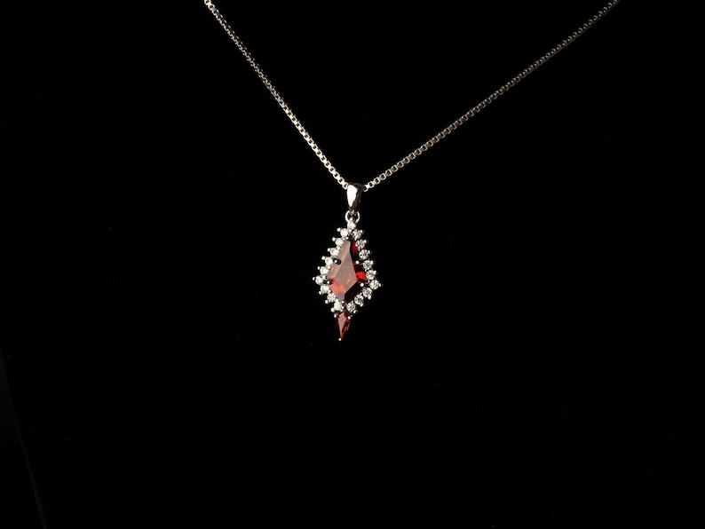 May include: A silver necklace with a pendant featuring a large, red gemstone surrounded by small, clear stones. The pendant has a unique, elongated shape. The background is solid black, highlighting the jewelry.