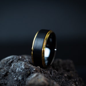 May include: A black tungsten carbide ring with a brushed finish and a gold band around the edge.