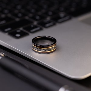 Peut inclure: Une bague noire et dor&eacute;e avec un incrustation en bois. La bague est pos&eacute;e sur un ordinateur portable argent&eacute;.
