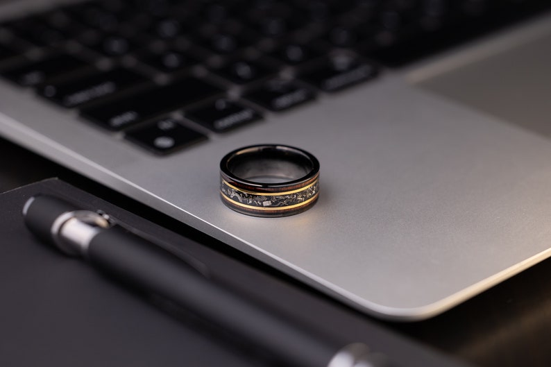 Peut inclure: Une bague noire et dor&eacute;e avec un incrustation en bois. La bague est pos&eacute;e sur un ordinateur portable argent&eacute;.