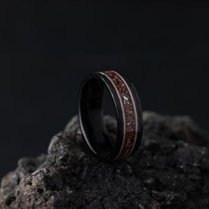 Puede incluir: Un anillo negro con un dise&ntilde;o de incrustaciones rojas y blancas. El anillo est&aacute; sobre una roca gris oscura.