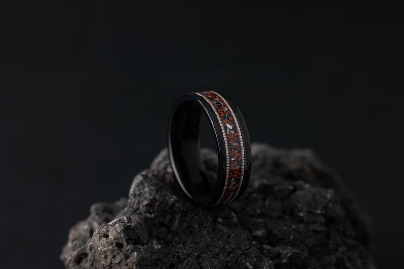 Puede incluir: Un anillo negro con un dise&ntilde;o de incrustaciones rojas y blancas. El anillo est&aacute; sobre una roca gris oscura.