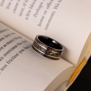 Peut inclure: Une bague en carbure de tungst&egrave;ne noir avec une incrustation brune et dor&eacute;e. La bague est pos&eacute;e sur un livre ouvert.