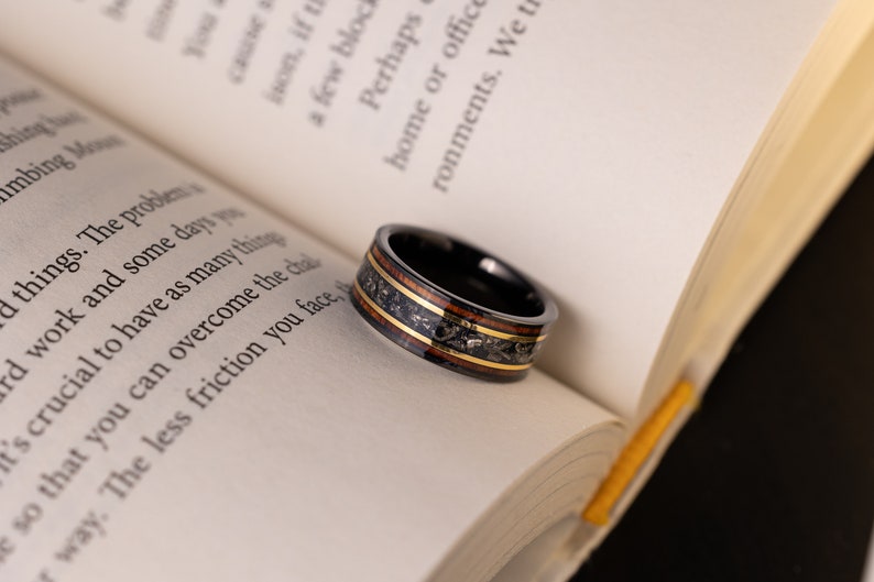 Peut inclure: Une bague en carbure de tungst&egrave;ne noir avec une incrustation brune et dor&eacute;e. La bague est pos&eacute;e sur un livre ouvert.