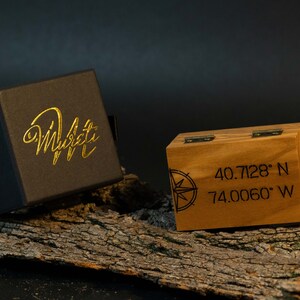 May include: A black box with gold lettering that reads "Murati" next to a wooden box with coordinates and a compass rose design. The wooden box is 8.9 cm wide and 6.4 cm tall.