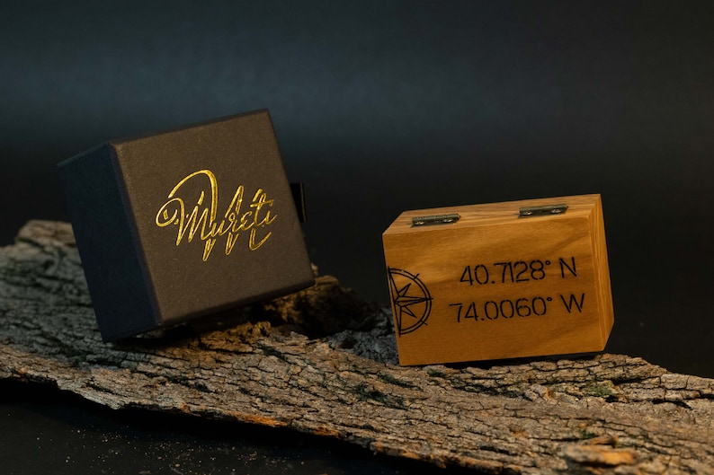 May include: A black box with gold lettering that reads "Murati" next to a wooden box with coordinates and a compass rose design. The wooden box is 8.9 cm wide and 6.4 cm tall.