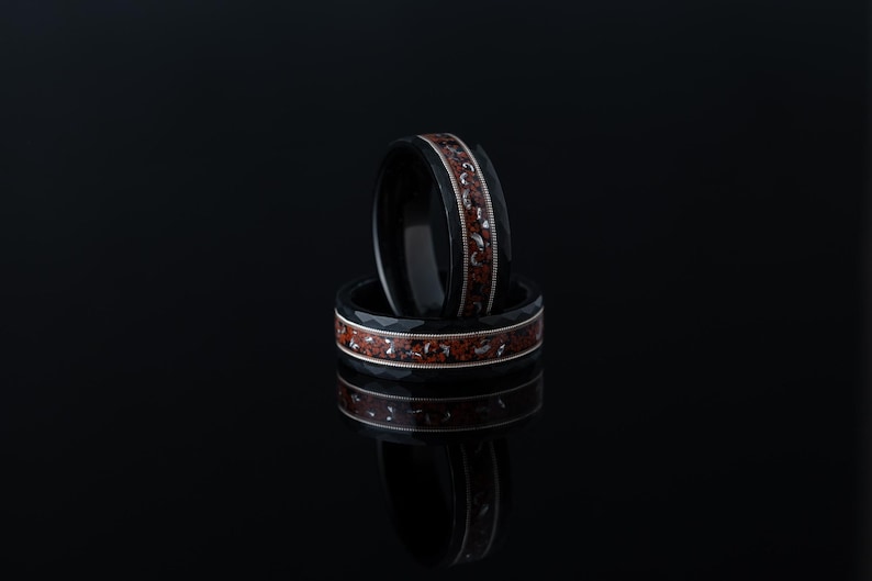 Puede incluir: Dos anillos negros con incrustaciones rojas y plateadas. Los anillos est&aacute;n apilados uno encima del otro.