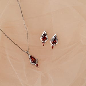 May include: A silver necklace and matching earrings set. The jewellery features a geometric design with a central, deep red gemstone surrounded by small, clear stones. The earrings and pendant have a similar shape.