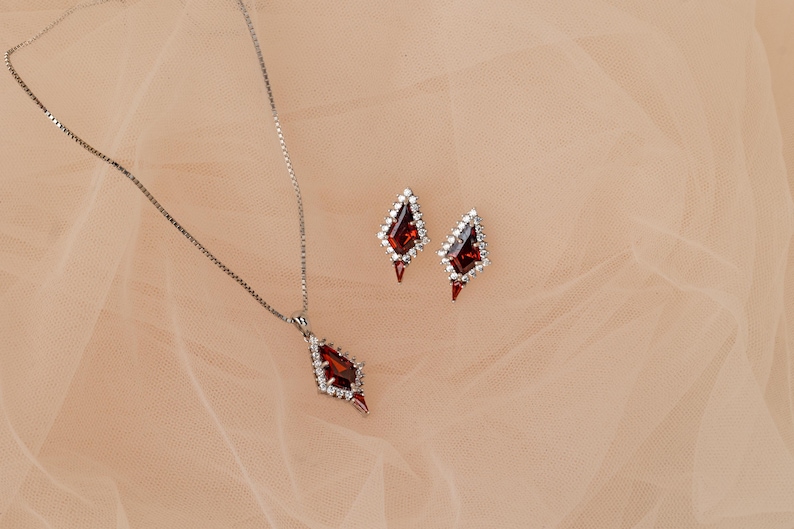 May include: A silver necklace and matching earrings set. The jewellery features a geometric design with a central, deep red gemstone surrounded by small, clear stones. The earrings and pendant have a similar shape.