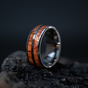 K&ouml;nnte beinhalten: Ein schwarzer Ring aus Wolframcarbid mit einer braunen Holzeinlage. Der Ring hat eine polierte Oberfl&auml;che und liegt auf einem dunklen Hintergrund.