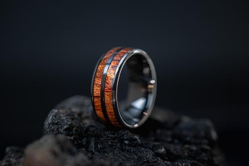 K&ouml;nnte beinhalten: Ein schwarzer Ring aus Wolframcarbid mit einer braunen Holzeinlage. Der Ring hat eine polierte Oberfl&auml;che und liegt auf einem dunklen Hintergrund.