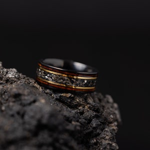Peut inclure: Une bague noire avec un incrustation dor&eacute;e et brune. La bague est pos&eacute;e sur une surface sombre et textur&eacute;e.