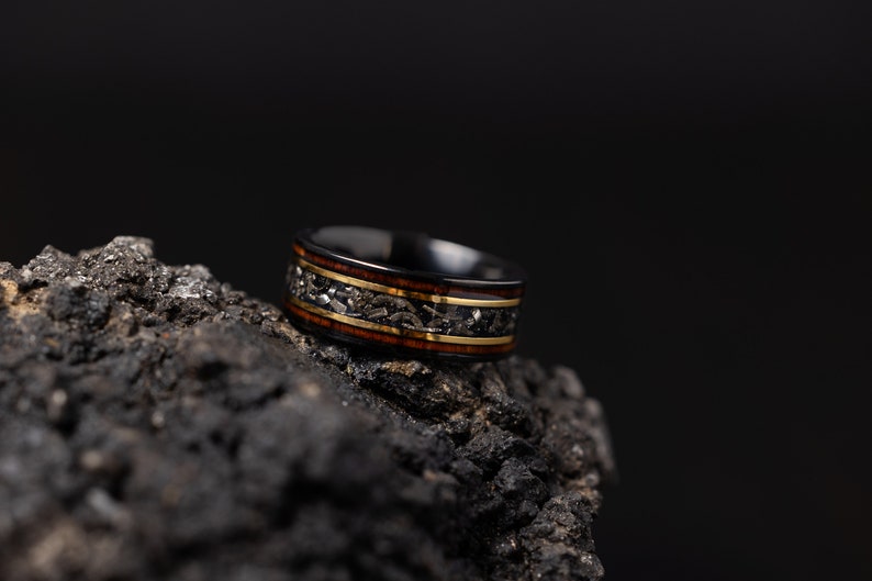Peut inclure: Une bague noire avec un incrustation dor&eacute;e et brune. La bague est pos&eacute;e sur une surface sombre et textur&eacute;e.