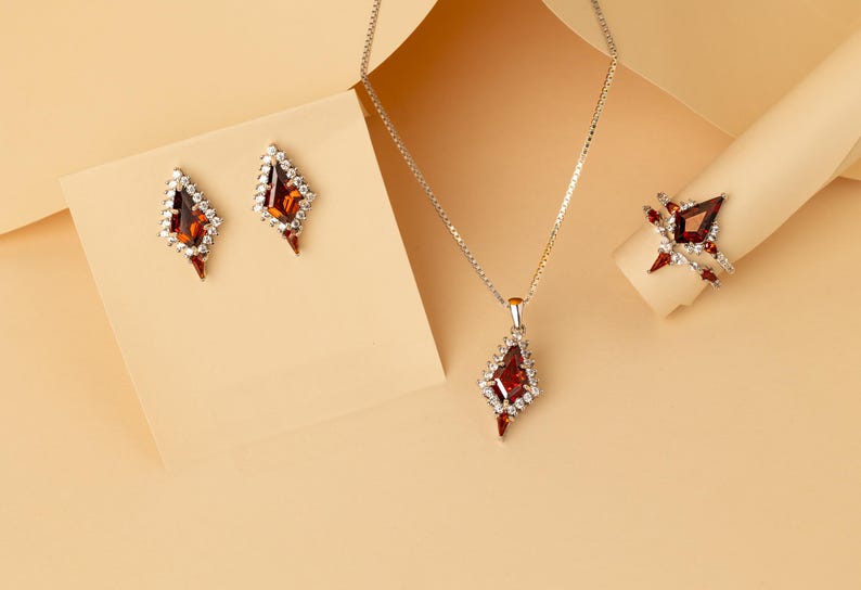 May include: A jewellery set comprising a necklace, earrings, and a ring. The pieces have a silver-coloured metal setting and are decorated with red and clear gemstones. The earrings and pendant are teardrop-shaped. The ring is on a beige surface.