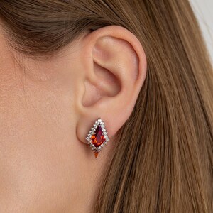 May include: A close-up of a diamond-shaped earring featuring a large, red gemstone at its centre. The gemstone is encircled by smaller, clear stones. The earring is set in silver or white metal and is worn on an ear.