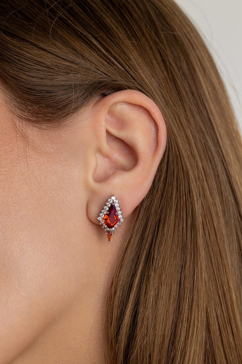 May include: A close-up of a diamond-shaped earring featuring a large, red gemstone at its centre. The gemstone is encircled by smaller, clear stones. The earring is set in silver or white metal and is worn on an ear.