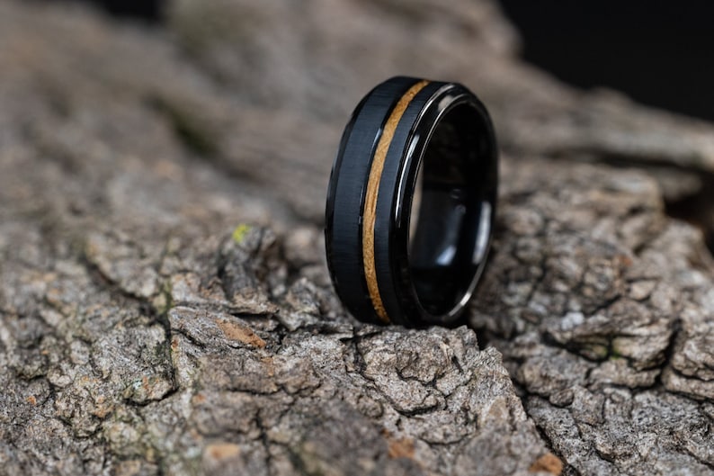 May include: A black tungsten carbide ring with a thin band of wood inlay. The ring is on a rough, textured surface.