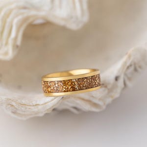 May include: A gold band ring with a textured surface of embedded gold flakes. The ring is resting on a white seashell.