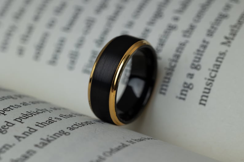 May include: A black and gold tungsten carbide ring with a brushed finish. The ring has a thin gold band around the edge.