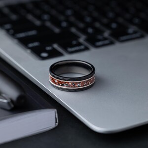 Puede incluir: Un anillo negro y plateado con una incrustaci&oacute;n roja. El anillo est&aacute; sobre un port&aacute;til plateado.