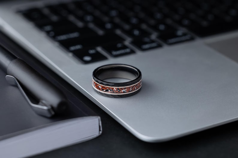 Puede incluir: Un anillo negro y plateado con una incrustaci&oacute;n roja. El anillo est&aacute; sobre un port&aacute;til plateado.