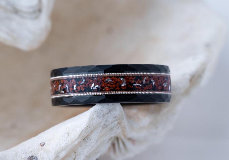 Puede incluir: Un anillo negro y plateado con una banda de incrustaciones rojas y plateadas. El anillo tiene un borde biselado y un acabado pulido.