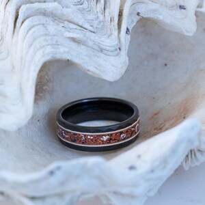 Puede incluir: Un anillo negro con un dise&ntilde;o de incrustaciones de cobre. El anillo est&aacute; sobre una concha blanca.