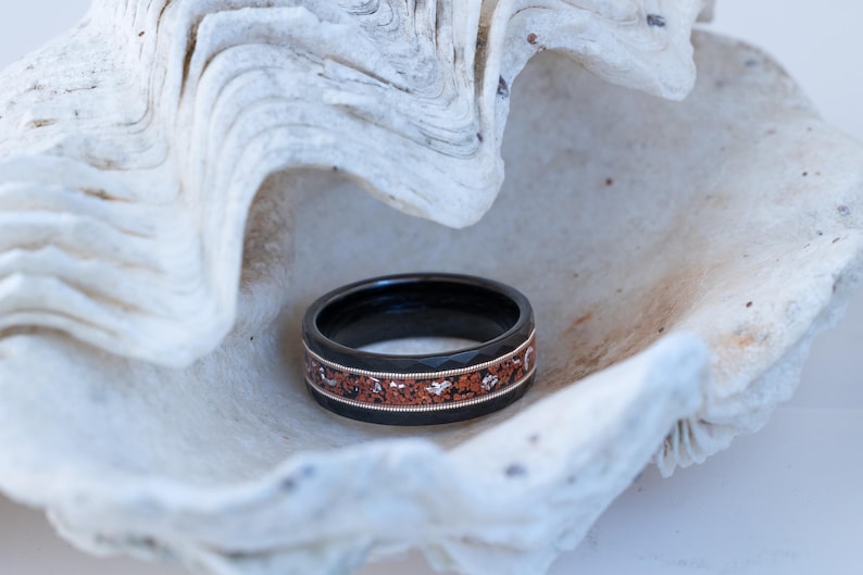 Puede incluir: Un anillo negro con un dise&ntilde;o de incrustaciones de cobre. El anillo est&aacute; sobre una concha blanca.