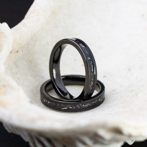 K&ouml;nnte beinhalten: Zwei schwarze Eheringe mit einer dezenten silbernen Einlage. Die Ringe sind &uuml;bereinander gestapelt und liegen in einer wei&szlig;en Muschel.