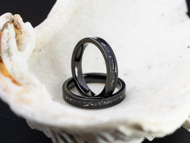 K&ouml;nnte beinhalten: Zwei schwarze Eheringe mit einer dezenten silbernen Einlage. Die Ringe sind &uuml;bereinander gestapelt und liegen in einer wei&szlig;en Muschel.