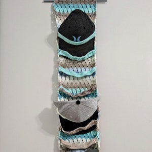 Può includere: Un'appendice da parete all'uncinetto con più tasche per riporre i cappelli. L'appendice è realizzata con filato beige, blu e bianco. Due cappelli sono visibili nelle tasche, uno grigio e uno nero.