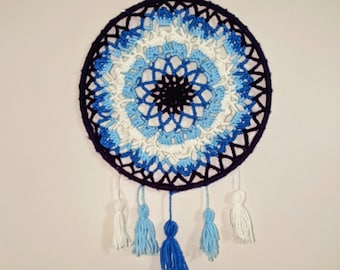 May include: A crocheted dream catcher with a purple, blue, and white color gradient. The dream catcher has a circular frame with a beaded center and white, blue, and purple tassels hanging from the bottom.