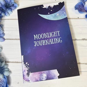 Peut inclure: Un journal rectangulaire avec une couverture sur le thème de la galaxie bleu et violet foncé, ornée d'un croissant de lune, d'étoiles et d'un motif de pleine lune. Le texte MOONLIGHT JOURNALING est imprimé en blanc au centre de la couverture.