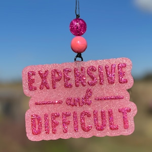 May include: A pink glittery car charm with the words "EXPENSIVE and DIFFICULT" in a playful font. The charm is suspended by a black string and features two decorative pink beads.