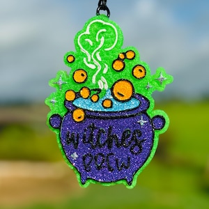 May include: A glittery, green, and purple Halloween-themed ornament. The ornament is shaped like a cauldron with the words "witches brew" in a purple glitter font. The cauldron has orange bubbles and green steam rising from it.