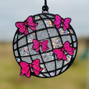 May include: A round, glittery ornament with a black grid pattern and bright pink bows. The ornament is filled with silver glitter and iridescent confetti. A black string is attached for hanging.