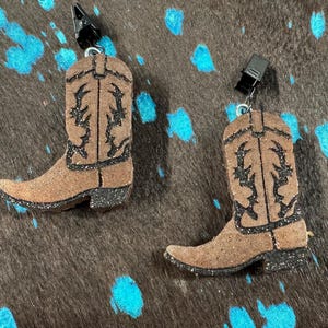May include: Two brown cowboy boot-shaped earrings with black glitter accents. The boots have a detailed design and are attached to black clips. The earrings are set against a brown and blue speckled background.