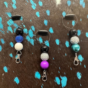 May include: Three beaded lanyards with black, white, blue, silver, purple, and teal beads. Each lanyard has a black loop, a series of beads, and a silver-colored clasp. The lanyards are displayed on a dark brown and blue patterned surface.
