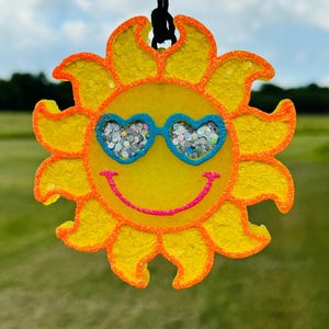 May include: A yellow sun-shaped ornament with orange trim and a black string for hanging. The sun has blue heart-shaped sunglasses filled with glitter and a pink smiling mouth. The background is a blurred green field and blue sky.