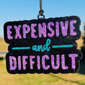 May include: Black glittery sign with the words "EXPENSIVE and DIFFICULT" in purple glitter lettering. The word "and" is in teal glitter. The sign is hanging from a black string.