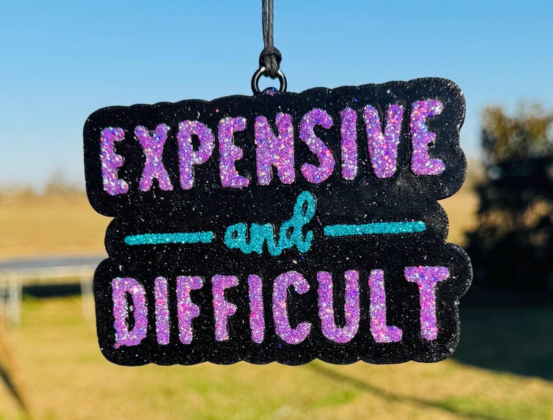 May include: Black glittery sign with the words "EXPENSIVE and DIFFICULT" in purple glitter lettering. The word "and" is in teal glitter. The sign is hanging from a black string.