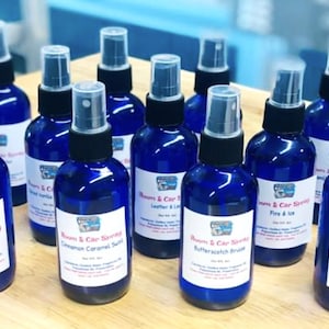 May include: A collection of ten blue glass spray bottles with white labels. The labels have the text "Room & Car Spraying" and different scents, including "Cinnamon Caramel Swirl", "Butterscotch Brulee", "Leather & Lace", and "Fire & Ice".